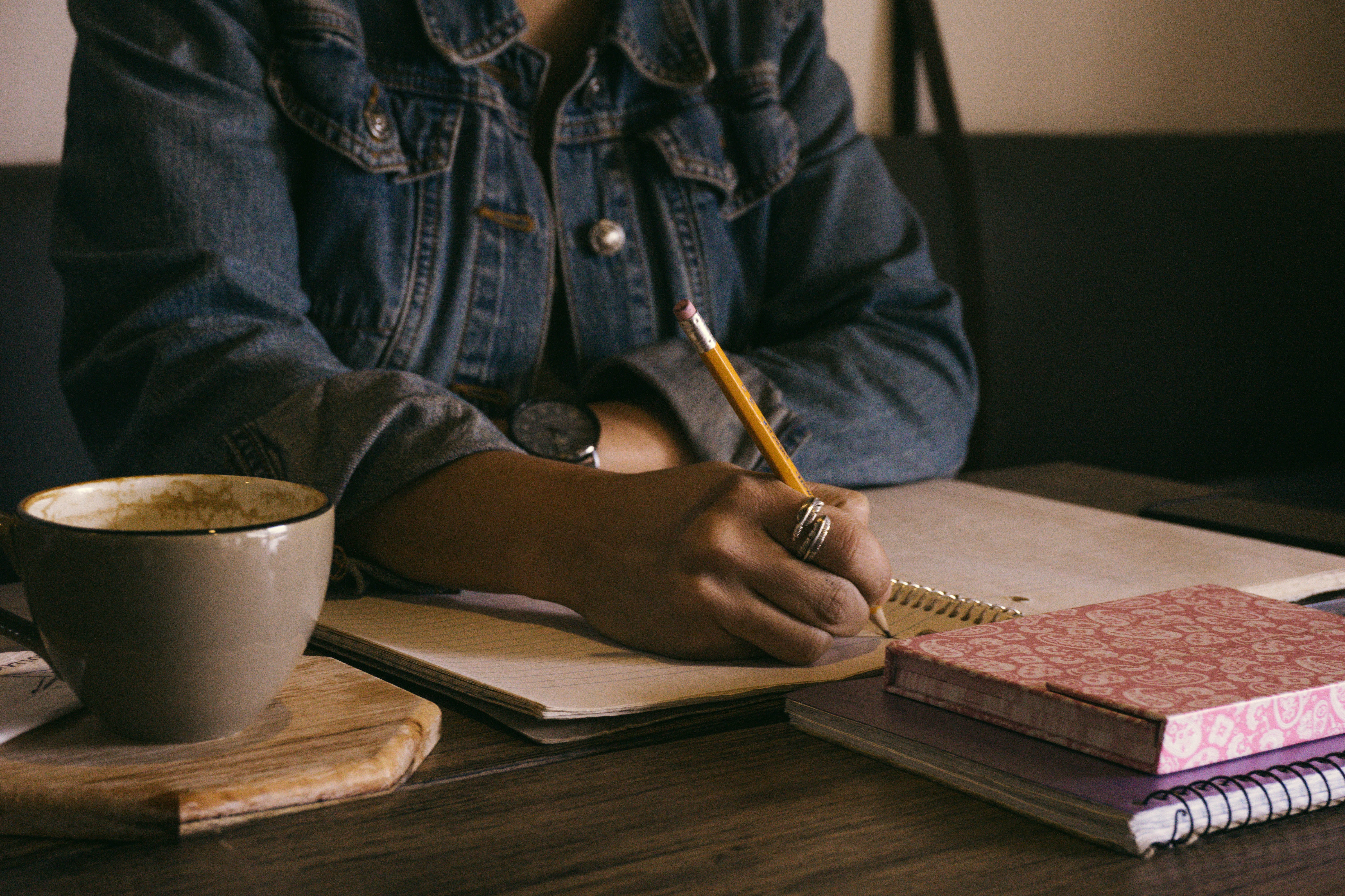 Person with ring on finger writing in a notepad beside a cup of coffee